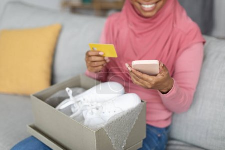 Téléchargez les photos : Delivery concept. Unrecognizable black muslim woman holding credit card and using smartphone, shopping online, sitting on couch with cardboard box container with new shoes - en image libre de droit