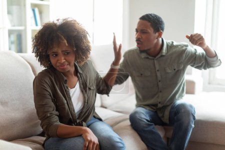 Foto de Angry furious young black man raising hand against his crying scared wife, african american couple have fight at home, spouses sitting on couch, arguing. Domestic violence, abuse, tyranny concept - Imagen libre de derechos