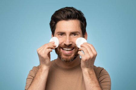 Téléchargez les photos : Portrait of happy bearded man holding cotton pads under eyes and smiling at camera, standing over blue studio background. Middle aged male cleansing face with toner - en image libre de droit