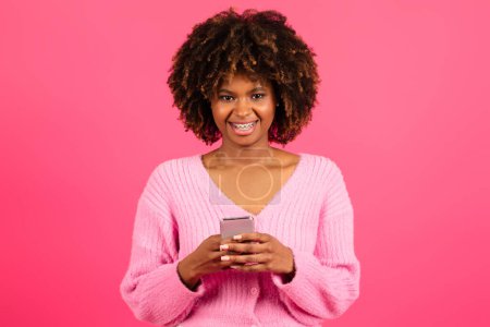 Foto de Cheerful young black curly lady in casual with braces typing on smartphone, read message, isolated on pink background, studio. App recommendation, surfing, blog and social media chat, ad and offer - Imagen libre de derechos