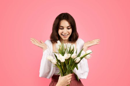 Téléchargez les photos : Glad surprised young caucasian female in casual rejoices gift with bouquet of flowers, spreads arms to sides isolated on pink background, studio. Emotions from celebration of holiday and birthday - en image libre de droit