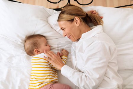 Téléchargez les photos : Loving mother hugging sleeping baby lying in bed in bedroom at home, top view. Infant girl sleeping near mom. Motherhood lifestyle, daytime sleep routine concept - en image libre de droit