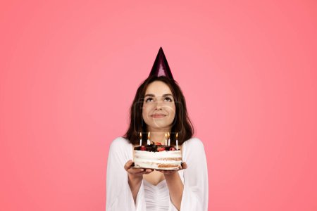 Photo for Happy pretty young caucasian female in hat hold cake with candles and looking up at empty space isolated on pink background, studio. Celebrating holiday, make wish for birthday and dream, ad and offer - Royalty Free Image
