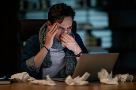 Téléchargez les photos : Sick male employee suffering from headache, working at office late at night, unhappy young man in casual with scarf around his neck sitting at workdesk, using computer, touching his head and sneezing - en image libre de droit