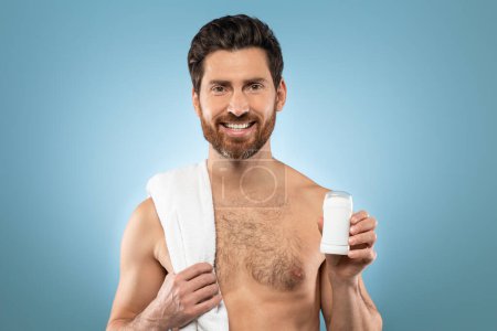 Photo for Positive shirtless caucasian man showing antiperspirant stick, standing with towel on shoulder over blue background. Male sweat protection, armpits skincare and hygiene - Royalty Free Image