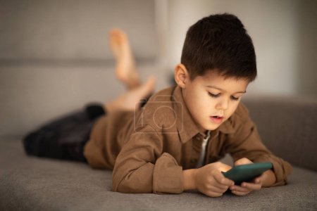 Busy cute small european boy lies on sofa, plays game, watches video on smartphone in room interior, close up, free space. Device for fun, video call and meeting remotely, app for learning at home