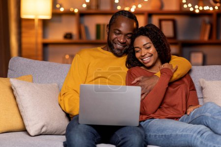 Foto de Happy smiling african american spouses sitting on couch in living room decorated with lights, bonding, using computer laptop, black man and woman shopping online from home, copy space - Imagen libre de derechos