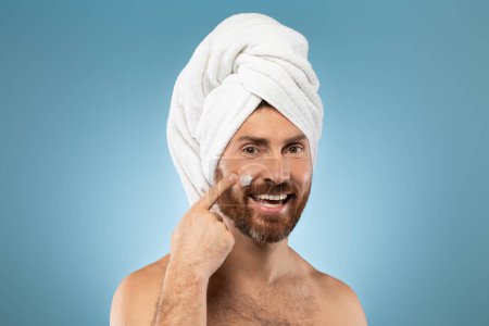 Photo for Mens skin care concept. Happy middle aged caucasian man smearing face cream on cheek and smiling at camera, posing on blue studio background, copy space - Royalty Free Image