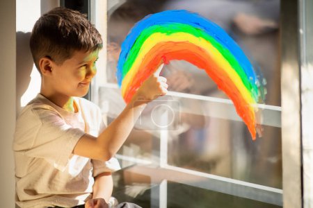 Téléchargez les photos : Cheerful small european boy draws rainbow with finger on window, enjoy spare time in children room interior, free space. Entertainment and fun, education, dreams, art and fantasy, childhood at home - en image libre de droit
