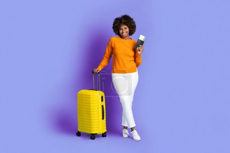 Téléchargez les photos : Cheery happy smiling pretty young black woman traveller carrying yellow luggage, holding passport and flight tickets, posing on purple studio background, copy space, full length. Tourism, traveling - en image libre de droit
