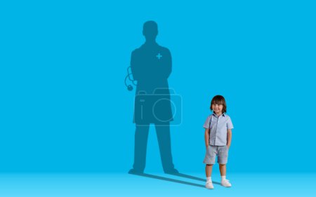 Foto de Childhood dreams concept. Cute handsome little boy dreaming to become a doctor, shadow of male doc behind kid posing over blue wall studio background, collage, copy space - Imagen libre de derechos