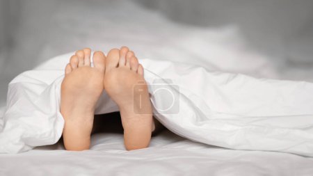 Téléchargez les photos : Legs of young european female lies on white comfortable bed under covers, wakes up, enjoys free time and vacation, panorama, close up, free space. Beauty and health care, rest and relaxation at home - en image libre de droit