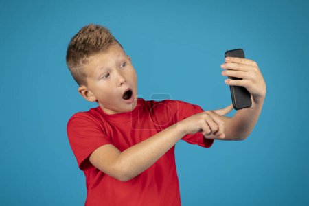 Téléchargez les photos : Portrait of scared preteen boy using smartphone and opening mouth in shock, frustrated male child reading message or watching photos on mobile phone, emotionally reacting to content, blue background - en image libre de droit
