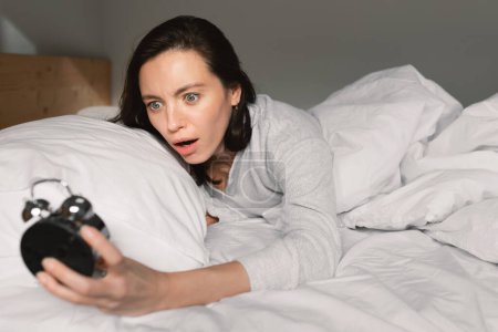 Foto de Shocked frightened young european woman lies on white comfortable bed, wake up, looks at alarm clock in bedroom interior, close up. Overslept, late for work, meeting at home and facial expressions - Imagen libre de derechos
