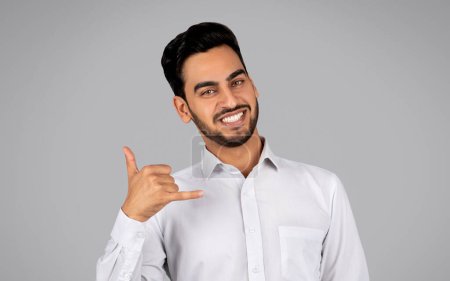 Foto de Call Me. Smiling Arab Businessman Making Phone Gesture And Looking At Camera, Handsome Middle Eastern Male Entrepreneur Inviting For Communication While Standing On Grey Background, Copy Space - Imagen libre de derechos