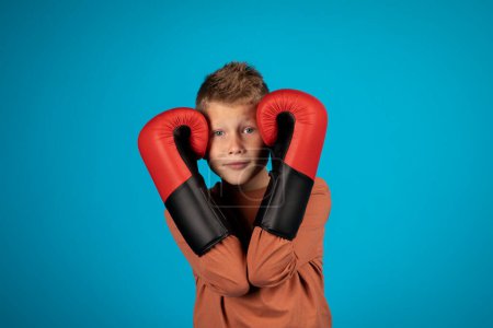 Photo for Portrait Of Preteen Blonde Boy Covering Ears With Boxing Gloves, Cute Male Child Protecting Himself From Punch And Looking At Camera, Standing Isolated Over Blue Studio Background, Copy Space - Royalty Free Image