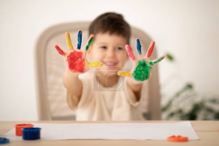 Photo for Cheerful small european boy with colorful hands palms enjoys drawing at table with paint in children room, school interior, blurred. Entertainment and fun, education, art and fantasy childhood at home - Royalty Free Image