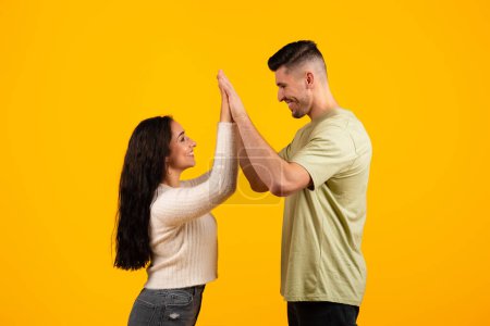 Téléchargez les photos : Cheerful young arabic woman and guy in casual, holding hands, doing high five, celebrating victory isolated on yellow background, studio, profile. Love, romance, relationship, success, ad and offer - en image libre de droit
