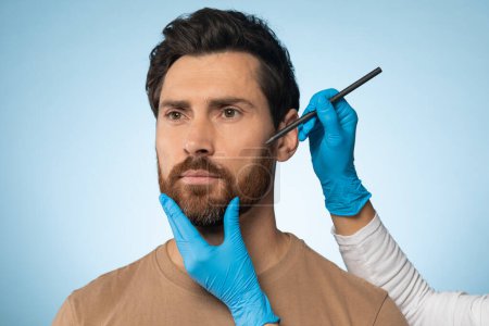 Téléchargez les photos : Aesthetic cosmetology concept. Man getting pencil marks on face skin for cosmetic surgery, plastic surgeon applying marks on male face before surgery, blue background - en image libre de droit