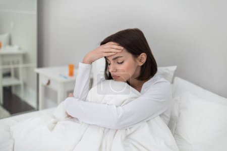 Foto de Sick young brunette woman sitting in bed under cover at home, touching head and hugging her knees, suffering from headache or fever while have cold or flue, copy space - Imagen libre de derechos
