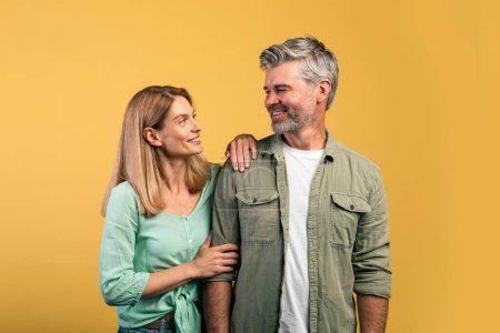 Foto de Positive middle aged caucasian spouses in casual outfits posing together, looking and smiling at each other over yellow studio background - Imagen libre de derechos
