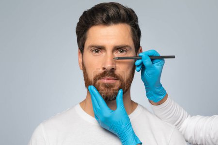 Doctor making marks on male patients face, middle aged man on consultation at surgeon, standing on grey background. Facial plastic surgery concept