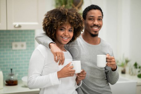 Téléchargez les photos : Portrait of sweet cheerful loving african american millennial couple embracing, cuddling, smiling at camera, chatting while drinking coffee at cozy kitchen in the morning, copy space - en image libre de droit