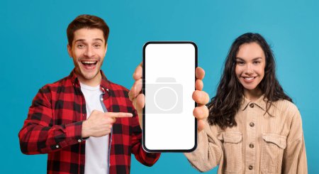 Téléchargez les photos : Mobile App. Joyful Young Man And Woman Pointing At Empty Cellphone With White Screen, Happy Millennial Couple Demonstrating Blank Smartphone With Copy Space For Advertisement Design, Mockup - en image libre de droit