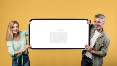 Foto de Mobile application ad. Happy mature couple holding big white empty smartphone screen, presenting cellphone display, mockup, yellow background - Imagen libre de derechos