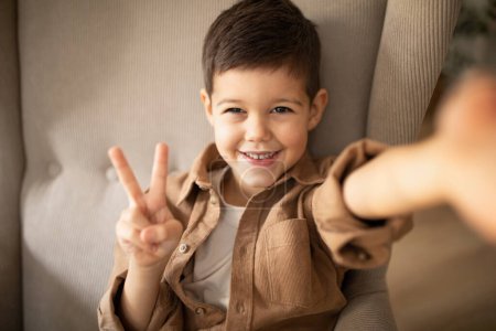 Photo for Smiling cute small european boy sitting on armchair, makes selfie and peace sign with hand in room interior, close up. Photo and device, video call and meeting remotely at home, fun and childhood - Royalty Free Image
