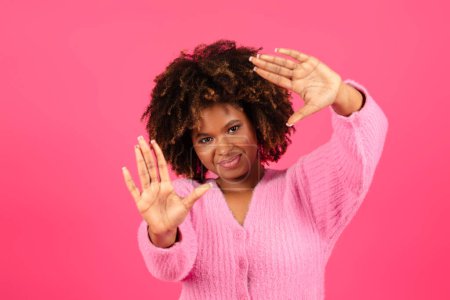 Téléchargez les photos : Cheerful young black curly lady in casual makes lens with hands, frame for shooting video, looks at imaginary camera, isolated on pink background. Photo for blog and social networks, offer and ad - en image libre de droit
