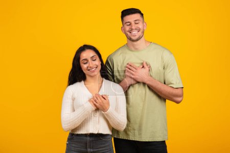 Téléchargez les photos : Cheerful relaxed young arabic woman and guy in casual with closed eyes put their hands to chest making gratitude sign, isolated on yellow background, studio. Thanks, wish and desire, ad and offer - en image libre de droit