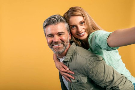 Foto de Having fun together. Happy middle aged spouses taking selfie, woman piggyback riding on her husbands back, looking at camera and smiling, yellow studio background, copy space - Imagen libre de derechos