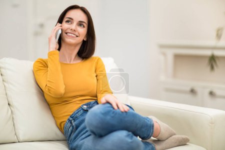 Foto de Happy cheerful smiling beautiful brunette young woman in comfy casual outwear sitting on couch with legs up, having phone conversation with friend or lover at home, copy space - Imagen libre de derechos