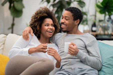 Téléchargez les photos : Cheerful loving millennial beautiful black man and woman reclining on couch in cozy living room decorated with green plants, drinking coffee, bonding, chatting and laughing, weekend at home concept - en image libre de droit