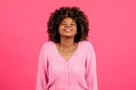 Photo for Satisfied young black curly lady with braces and closed eyes inhales deeply, enjoys freedom, raises her head, isolated on pink background, studio. Emotions from rest and relax, freedom and lifestyle - Royalty Free Image