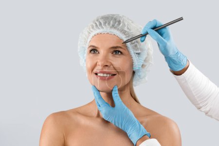 Téléchargez les photos : Plastic surgery or face lifting for middle aged woman. Doctor hands wearing gloves drawing lines, holding head of female patient, isolated on grey background, copy space - en image libre de droit