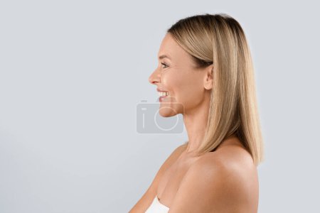 Photo for Profile portrait of middle aged blonde woman isolated on grey, smiling beautiful adult lady wrapped in white towel demonstrating perfect face contours, looking at copy space. Injection cosmetology - Royalty Free Image