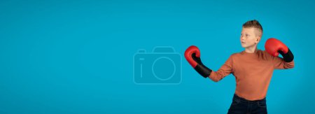 Téléchargez les photos : Preteen Boy Wearing Boxing Gloves Making Fighter Posture And Looking Aside At Copy Space, Motivated Male Kid Ready To Make Punch, Standing Isolated Over Blue Studio Background, Panorama - en image libre de droit