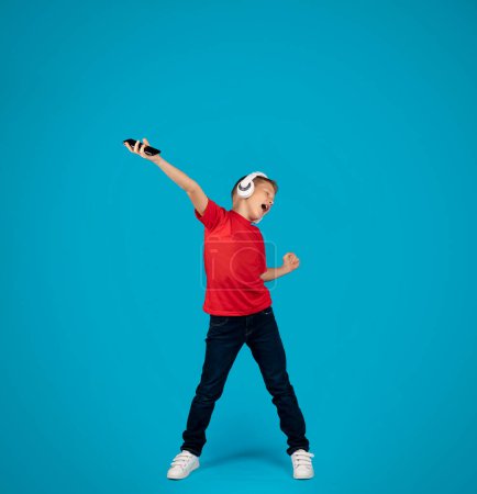 Téléchargez les photos : Cheerful preteen boy in wireless headphones listening music on smartphone and dancing, positive male child enjoying favorite songs, having fun while standing on blue studio background, copy space - en image libre de droit