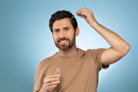 Téléchargez les photos : Happy handsome caucasian man applying anti-aging serum and smiling at camera, posing over blue studio background. Cheerful male using nourishing serum for his face - en image libre de droit