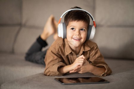 Foto de Thoughtful happy small european boy in headphones lies on sofa, thinks, dreams with tablet, watch videos and play games, look at free space in room interior. Study at home with device and free time - Imagen libre de derechos