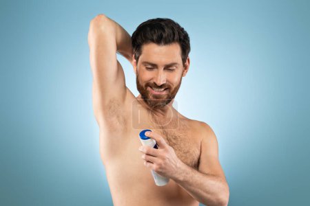 Photo for Shirtless caucasian man putting deodorant underarm after shower, standing over blue background, studio shot, free space. Handsome male doing morning routine - Royalty Free Image