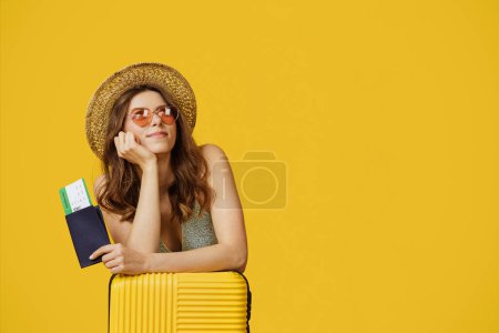 Foto de Happy female traveler holding passport and travel tickets, sitting with suitcase and dreaming, looking aside over yellow background, free space. Vacation and tourism concept - Imagen libre de derechos