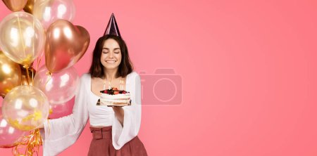 Cheerful young caucasian woman in hat hold a lot of inflatable balloons, blows out candles on cake isolated on pink background, studio. Emotions from celebrating holiday and make wish for birthday
