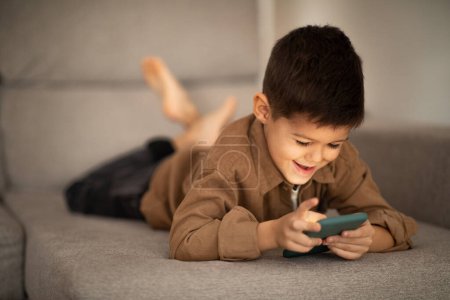 Foto de Busy cheerful small european boy lies on sofa, plays game, watches video on smartphone in room interior, free space. Gadget, video call and meeting remotely, app for learning and spare time at home - Imagen libre de derechos
