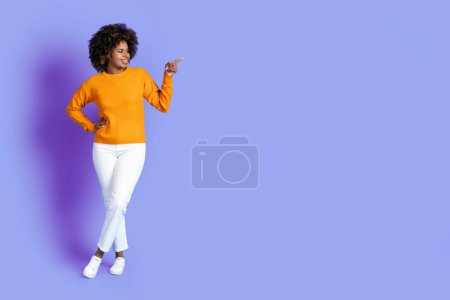 Téléchargez les photos : Nice offer. Positive cheerful stylish pretty curly young black woman in casual outfit pointing at copy space for ad over purple studio background, full length, panorama - en image libre de droit