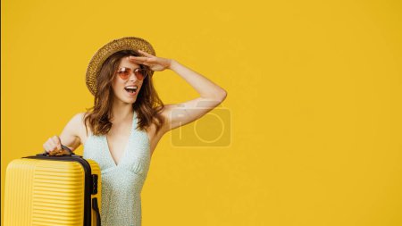 Téléchargez les photos : Happy young woman with straw hat and sunglasses carrying suitcase and looking aside at copy space over yellow studio background, panorama - en image libre de droit