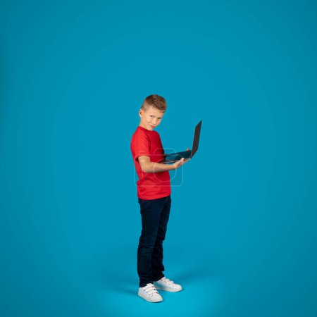 Foto de Full Length Shot Of Cute Preteen Boy Holding Laptop Computer And Looking At Camera While Standing Over Blue Background In Studio, Smiling Male Child Study Online Or Playing Video Games, Copy Space - Imagen libre de derechos