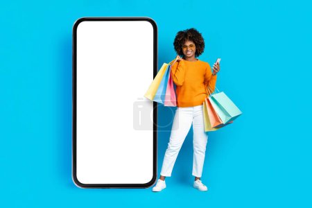 Photo for Cool stylish happy smiling young black woman with bushy hair holding colorful shopping bags and smartphone, wearing sunglasses, posing by big phone with mockup over blue studio background. E-commerce - Royalty Free Image